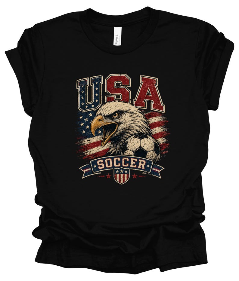 Peut inclure: Un t-shirt noir &agrave; manches courtes avec une impression graphique repr&eacute;sentant un pygargue &agrave; t&ecirc;te blanche, un ballon de football et le drapeau am&eacute;ricain. Le texte USA est imprim&eacute; en grandes lettres au-dessus de l'aigle et SOCCER sur une banni&egrave;re.