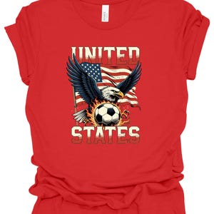 Op de afbeelding: Een rood t-shirt met korte mouwen en een afbeelding van een Amerikaanse zeearend met gespreide vleugels boven een voetbal, tegen een wapperende Amerikaanse vlag. De tekst UNITED staat boven de adelaar en STATES onder de voetbal.