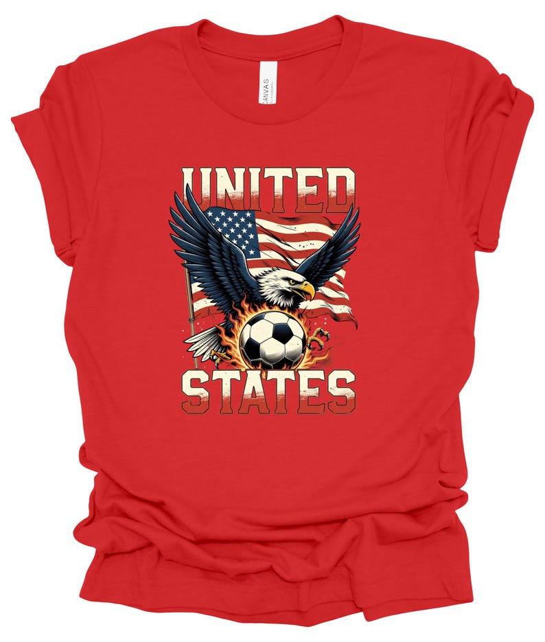 Op de afbeelding: Een rood t-shirt met korte mouwen en een afbeelding van een Amerikaanse zeearend met gespreide vleugels boven een voetbal, tegen een wapperende Amerikaanse vlag. De tekst UNITED staat boven de adelaar en STATES onder de voetbal.