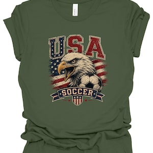 Peut inclure: Un t-shirt vert olive avec une impression graphique repr&eacute;sentant un pygargue &agrave; t&ecirc;te blanche, un ballon de football et le drapeau am&eacute;ricain. Le texte "USA" est en rouge et bleu au-dessus, et "SOCCER" est sur une banni&egrave;re bleue en dessous.
