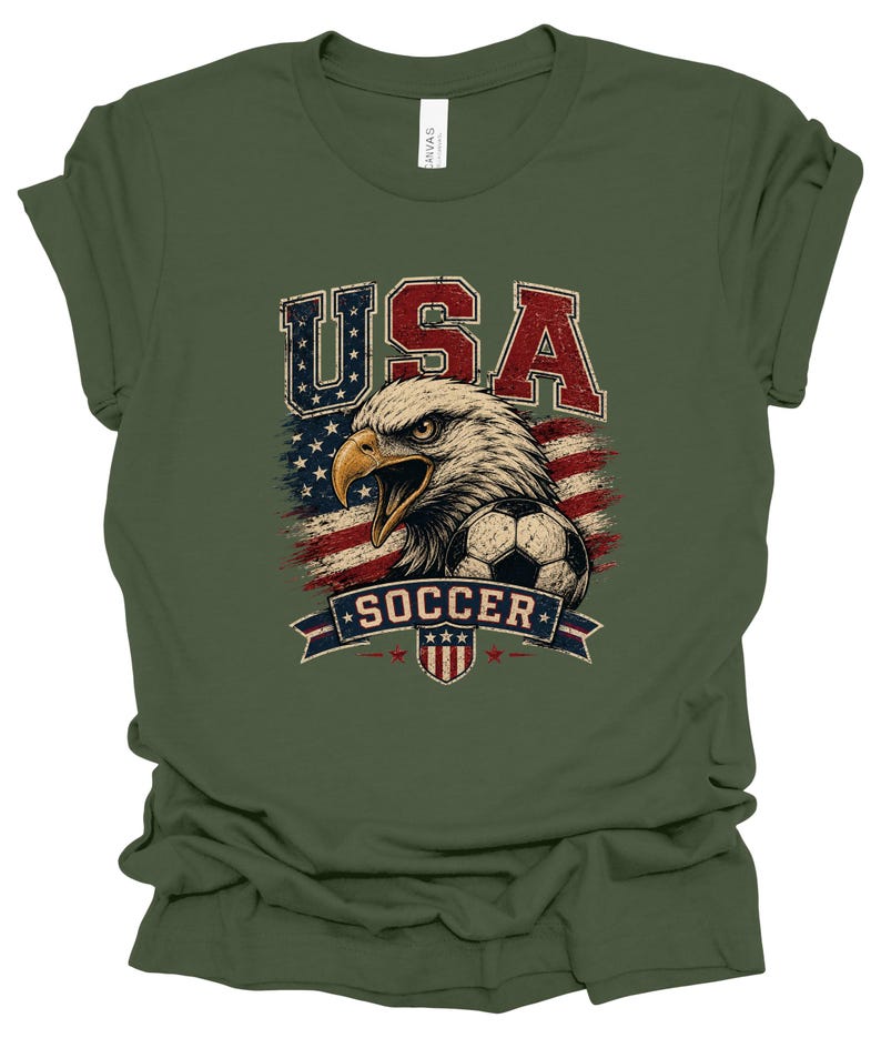 Peut inclure: Un t-shirt vert olive avec une impression graphique repr&eacute;sentant un pygargue &agrave; t&ecirc;te blanche, un ballon de football et le drapeau am&eacute;ricain. Le texte "USA" est en rouge et bleu au-dessus, et "SOCCER" est sur une banni&egrave;re bleue en dessous.