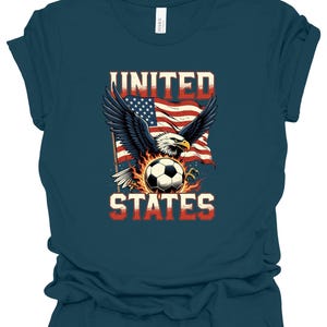 Op de afbeelding: Een donkerblauwgroen t-shirt met een afbeelding van een Amerikaanse zeearend met gespreide vleugels boven een voetbal omringd door vlammen. Achter de adelaar wappert een Amerikaanse vlag. De tekst UNITED staat boven de adelaar en STATES eronder.