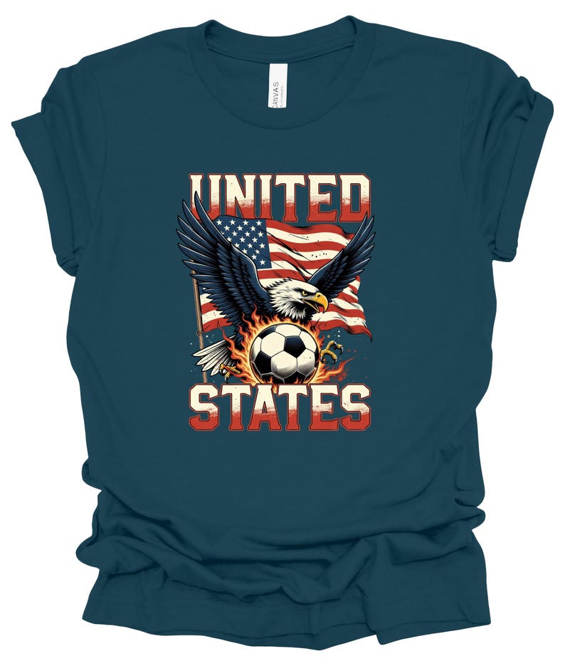 Op de afbeelding: Een donkerblauwgroen t-shirt met een afbeelding van een Amerikaanse zeearend met gespreide vleugels boven een voetbal omringd door vlammen. Achter de adelaar wappert een Amerikaanse vlag. De tekst UNITED staat boven de adelaar en STATES eronder.