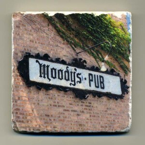 May include: A square coaster featuring a brick wall with green vines and a sign that reads "Moody's PUB" in black gothic lettering. The sign has a black decorative border and a small light fixture.