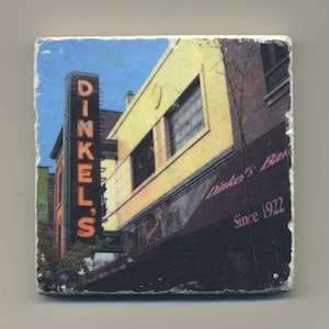 May include: Square coaster featuring a vintage photograph of Dinkel's Bakery. The image shows a neon sign that reads "Dinkel's" and the bakery's name on the awning, with "Since 1922" below. The building is yellow and brick.