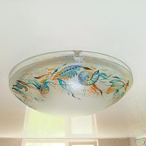 May include: A circular, frosted glass flush mount ceiling light fixture featuring a hand-painted marine design. The artwork displays colorful seashells, including a prominent nautilus, alongside seaweed and coral in shades of blue, orange, and green.