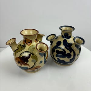 May include: Two small ceramic bud vases with four openings each. One vase has a floral design in shades of brown, blue, and green on a cream background. The other vase has a blue and white swirling design on a cream background.