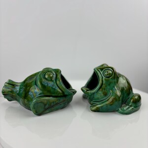 May include: Two ceramic frog ornaments with a glossy, green glaze. The frogs have open mouths and are positioned side-by-side. These are likely decorative items or small containers.