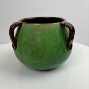 May include: A small green ceramic pot with two brown handles. The pot has a rounded shape and a smooth, glossy finish.