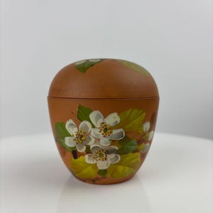 Può includere: Un piccolo contenitore con coperchio color terracotta con un motivo floreale dipinto a mano. I fiori sono bianchi con centri gialli e foglie verdi. Il contenitore ha una forma arrotondata e poggia su una superficie bianca.