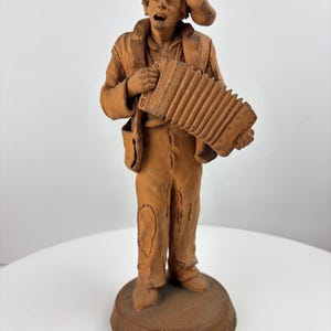 Può includere: Statuetta in terracotta di un musicista che suona la fisarmonica. La figura indossa un cappello e una giacca, con una rappresentazione dettagliata dello strumento. La statua è su una base rotonda, mostrando un'estetica rustica e artigianale.