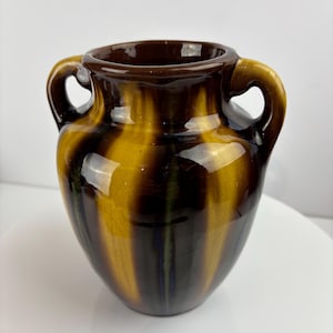 Puede incluir: Un jarrón de cerámica con dos asas, con un acabado brillante. El jarrón tiene rayas verticales en tonos marrones y amarillos. El interior y el borde son de color marrón oscuro.
