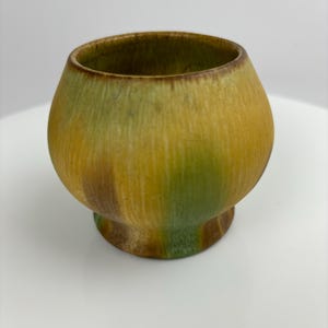 May include: A small, round ceramic bowl with a green, yellow and brown glaze. The bowl has a slightly flared rim and a rounded base.