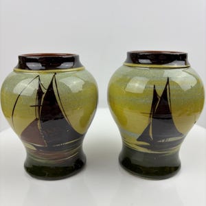 May include: Two ceramic jars with a sailboat design. The jars have a rounded shape with a dark brown rim and base. The body of the jars is a mix of green and yellow, with a sailboat illustration in dark brown.