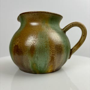May include: A brown and green ceramic jug with a rounded body and a handle. The jug has a mottled glaze with shades of brown, green, and yellow.