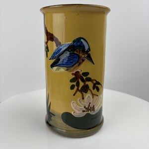 Può includere: Un vaso cilindrico in ceramica con sfondo giallo. Il vaso presenta un disegno dipinto a mano di un uccello blu e nero appollaiato su un ramo, con un fiore bianco e foglie verdi alla base. Il vaso ha un bordo leggermente svasato.
