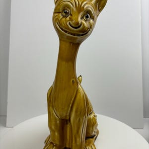 May include: A ceramic cat figurine with a brown glaze. The cat has a wide, toothy grin and large, round eyes. The cat is sitting upright with its tail curled around its legs.