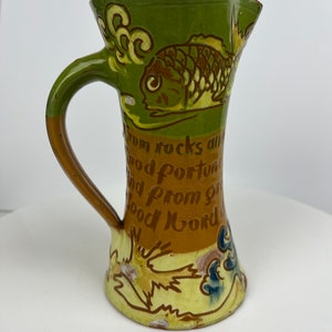 Może przedstawiać: Wazon ceramiczny w stylu vintage w kolorach zielonym, brązowym i żółtym. Posiada wzór ryby i napis: "From rocks and good fortune and from good word".