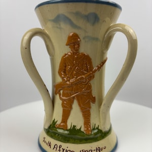 Könnte beinhalten: Eine Keramikvase mit zwei Henkeln, die einen Soldaten in brauner Uniform mit einem Gewehr darstellt. Die Vase ist mit blauen und grünen Akzenten verziert und trägt den Text "South Africa 1899-1900" auf dem Boden.