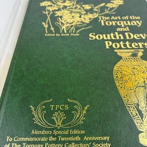 May include: Green book cover with gold lettering that reads "The Art of the Torquay and South Devon Potters". The cover also features a gold illustration of a vase and a floral design. The text "Members Special Edition" and "To Commemorate the Twentieth Anniversary of The Torquay Pottery Collectors' Society" is also printed in gold.