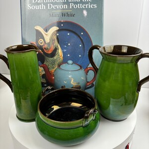 以下が含まれることがあります： 緑と茶色の陶器のセットで、ピッチャー、花瓶、ボウル、ティーポットが含まれています。背景にはマット・ホワイト著の「Dartmouth and the South Devon Potteries」という本が見えます。