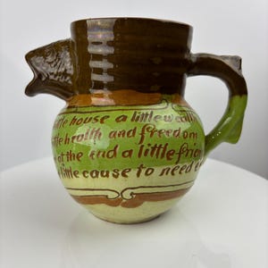 May include: A brown and green ceramic pitcher with a fish head spout and a handle shaped like a branch. The pitcher has a quote written on it: "A little house, a little wealth, a little health and freedom, at the end a little friend, a little cause to need em."