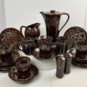 Puede incluir: Un juego de tazas de té, platillos y una tetera de cerámica moteada en marrón y blanco. El juego incluye una jarra de crema y un azucarero.