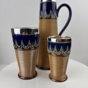 May include: A vintage ceramic jug and two matching glasses. The jug and glasses have a brown ribbed body, a dark blue top with a silver rim, and a decorative pattern. The jug has a handle and a silver lid.