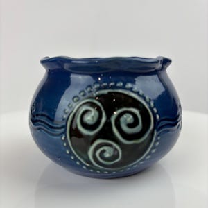Könnte beinhalten: Eine kleine, runde, blaue Keramikschale mit gewelltem Rand. Die Schale hat ein schwarz-weißes keltisches Spiraldesign auf der Vorderseite, mit zusätzlichen dekorativen Linien und Punkten. Die Schale ist wahrscheinlich handgefertigt.