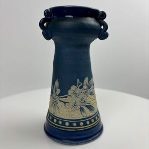 May include: A dark blue ceramic vase with a flared base and a narrow neck. The vase features a floral design in a lighter colour around the middle and decorative handles near the top. The vase is approximately 15 cm tall.
