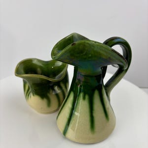 Op de afbeelding: Twee kleine keramische kannen met groene en witte glazuur. De kannen hebben een unieke, licht asymmetrische vorm.