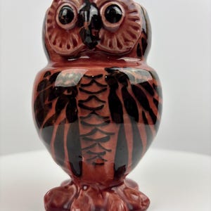 Puede incluir: Figura de búho de cerámica en tonos marrón y negro. El búho tiene ojos grandes y oscuros y un patrón de plumas detallado. La figura se asienta sobre una base que se asemeja a raíces de árbol, creando una pieza decorativa caprichosa.
