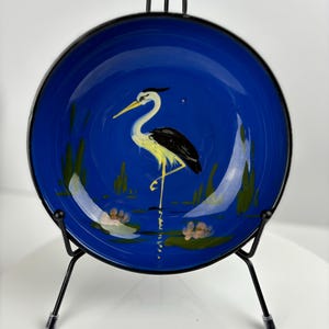 Könnte beinhalten: Ein blauer Keramikteller mit einem handgemalten Bild eines gelben und schwarzen Reihers, der in einem Teich mit grünen Schilf und rosa Blumen steht. Der Teller steht auf einem schwarzen Metallständer.