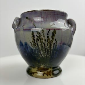 Può includere: Un piccolo vaso in ceramica dipinto a mano con due manici. Il vaso è decorato con un motivo floreale in tonalità di blu, verde e viola.
