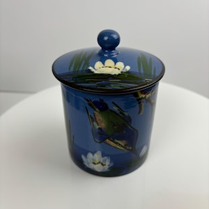 May include: A blue ceramic jar with a lid, decorated with a painted scene of birds and water lilies. The jar has a dark blue glaze and the lid has a knob.