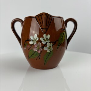 May include: A brown ceramic jug with two handles and a floral design painted on the side. The flowers are white with pink centres and green leaves.