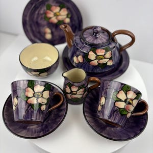 May include: A purple and white floral tea set with a teapot, creamer, two cups and saucers, and a plate. The set is hand-painted with pink flowers and green leaves.