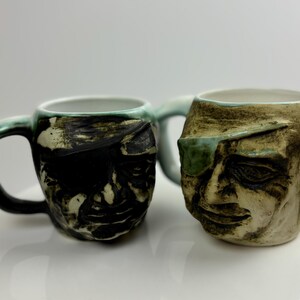 Puede incluir: Dos tazas de cerámica con diseños faciales únicos. Una taza tiene una cara verde y negra con un parche en el ojo negro. La otra taza tiene una cara verde y marrón con un parche en el ojo verde.