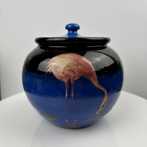 May include: A blue and black ceramic jar with a lid, featuring a hand-painted pink flamingo on a blue background.
