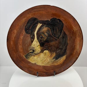 May include: A circular, wooden decorative plate with a painted portrait of a dog. The dog has black and brown fur with a white stripe down its face and a tan muzzle. The plate has a brown border.