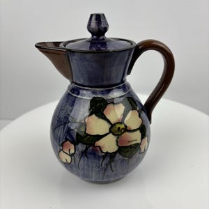Peut inclure: Un petit pichet en céramique bleu avec une anse et un couvercle marrons. Le pichet est décoré d'une fleur blanche avec des accents roses et des feuilles vertes.