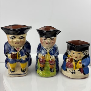 Puede incluir: Tres figuras de cerámica que representan a hombres con vestimenta tradicional. Cada figura tiene un borde marrón, sombrero negro y abrigo azul. Las figuras sostienen tazas y tienen pantalones amarillos. Las figuras están sobre una superficie blanca.