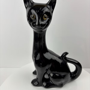 May include: A glossy black ceramic cat figurine with a long neck, pointed ears, and a curled tail. The cat has yellow eyes and is sitting upright. The figurine has a vintage aesthetic.