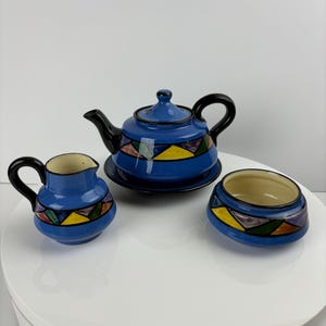 Op de afbeelding: Een blauwe keramische theepot, roomkan en suikerpot set met een geometrisch patroon in geel, groen, paars en zwart.
