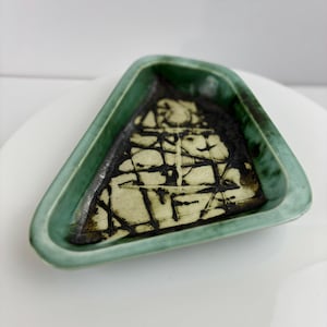 Puede incluir: Un plato triangular de cerámica con borde verde e interior texturizado. El interior presenta una superficie color crema con un patrón negro en forma de rejilla. El plato es probablemente hecho a mano y adecuado para objetos pequeños.
