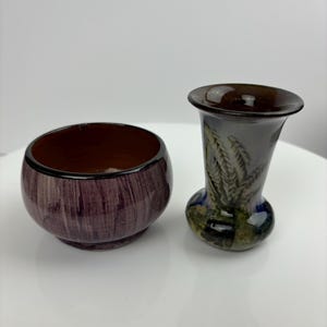 Puede incluir: Dos jarrones de cerámica, uno un tazón pequeño con un esmalte marrón y morado y el otro un jarrón más alto con un diseño de hojas en tonos de verde, azul y marrón.