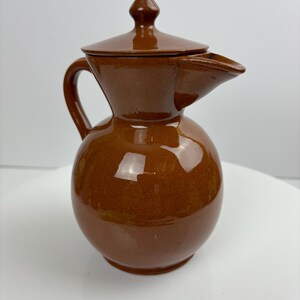 May include: A brown ceramic jug with a lid and a spout. The jug has a rounded body, a handle, and a lid with a small knob. The surface is glossy and reflects light. The jug is likely used for pouring liquids.