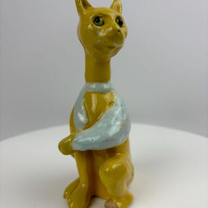 May include: A yellow ceramic cat figurine with a blue and white collar. The cat is sitting with its front paws crossed and has green eyes.
