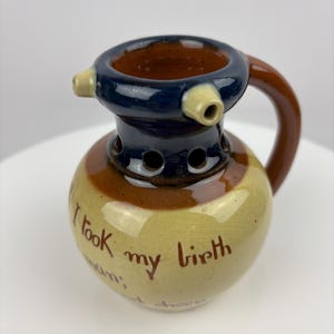 Puede incluir: Una pequeña jarra de cerámica marrón y amarilla con una parte superior azul y un asa. La jarra tiene un pico y agujeros alrededor de la parte superior. La jarra tiene el texto "I took my birth" escrito en ella.