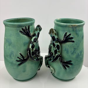 May include: A pair of green ceramic vases, each adorned with a frog motif. The vases have a cylindrical form with a subtle flared top. The frogs feature black and green spots and are attached to the front.